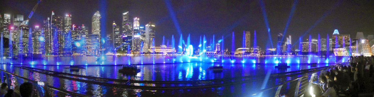 Night panorama of Singapore Marina Bay showing illuminated skyscrapers, blue light beams, and water fountain show with spectators along the waterfront