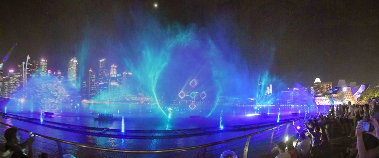 Night panorama of Singapore's Marina Bay Spectra light and water show with blue and green projections on water fountains, CBD skyline in background, and crowds of spectators watching from the waterfront promenade