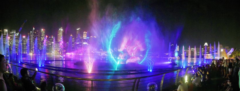 Night view of Singapore Marina Bay light and water show with colorful purple and blue fountain projections, illuminated skyscrapers in background, and spectators watching from waterfront promenade