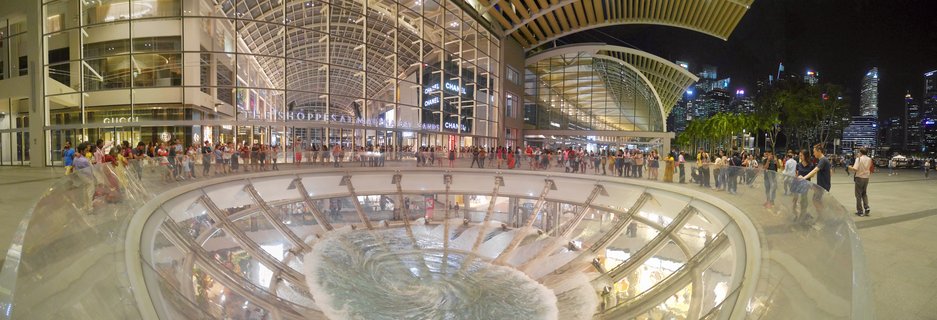 Night photograph showing the Rain Oculus water feature at Marina Bay Sands shopping mall in Singapore, with crowds of tourists around the circular glass structure and luxury brand storefronts visible in the background