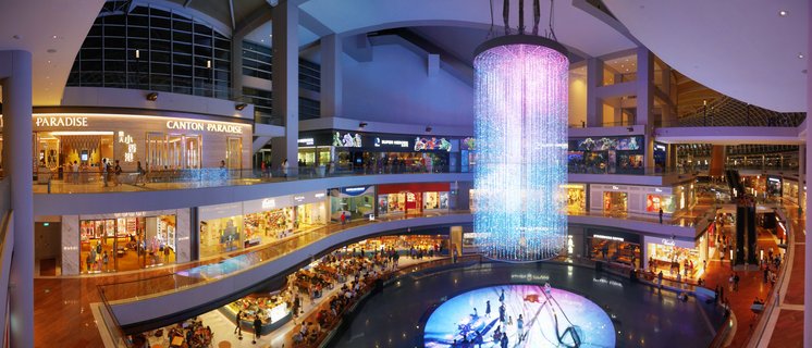 Multi-level luxury shopping mall interior with large cylindrical crystal chandelier, visible store signs including Canton Paradise and Starbucks, shoppers and diners on multiple floors