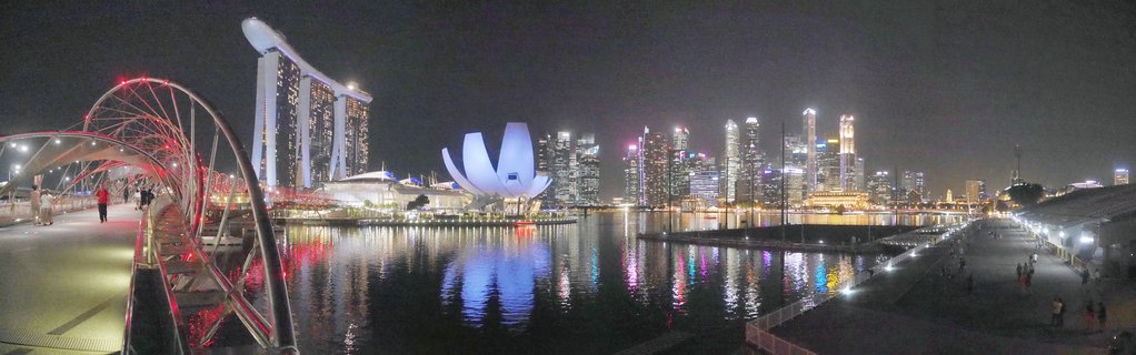Panoramic night photograph of Singapore Marina Bay showing illuminated Marina Bay Sands hotel, ArtScience Museum, CBD skyscrapers, and Helix Bridge with pedestrians, all reflected in dark water
