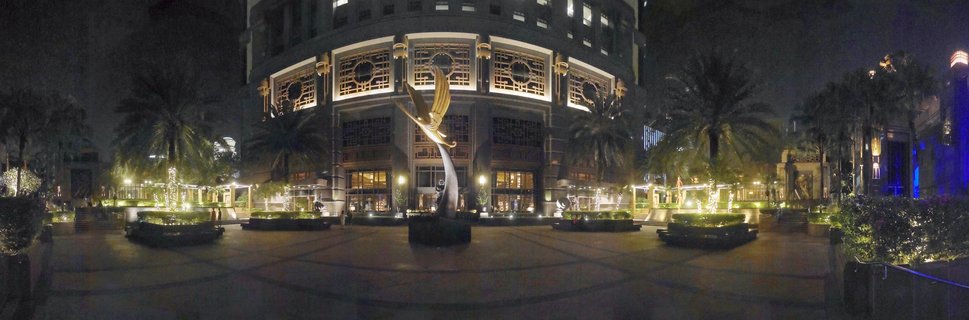 Night photograph of a luxury hotel plaza in Singapore with a golden winged sculpture in the center, surrounded by palm trees and an illuminated building with ornate window grilles