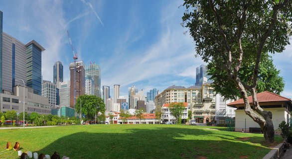 Panoramic cityscape showing Singapore's modern skyscrapers behind colonial buildings with red roofs, viewed across a large green lawn with trees under a blue sky with wispy clouds