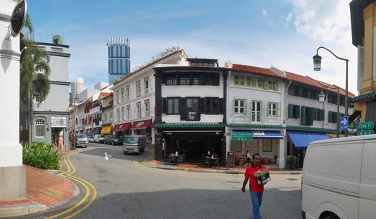 Street view of colorful two-story shophouses with red tile roofs at a Singapore street corner, with a pedestrian in red shirt walking past and a modern skyscraper visible in the distance under a blue sky
