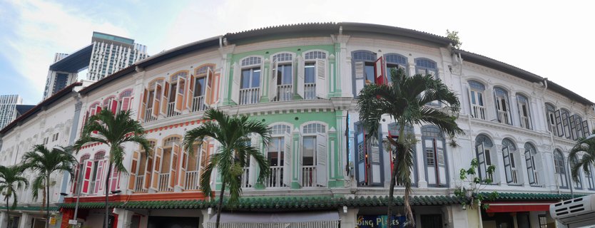 Street-level view of colorful two-story shophouses in pastel green, orange, white and blue with traditional shutters and palm trees in front, modern buildings visible in background