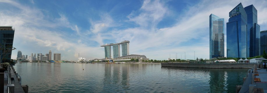 Panoramic view of Singapore's Marina Bay showing the Marina Bay Sands hotel with its distinctive rooftop sky park, surrounded by modern glass skyscrapers reflected in the bay water under a blue sky with wispy clouds