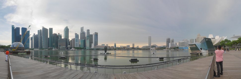Panoramic cityscape showing Singapore's Marina Bay with modern skyscrapers, the ArtScience Museum, calm water with floating barriers, and a wooden boardwalk with people in the foreground under cloudy sky