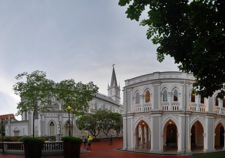 White Gothic Revival cathedral with tall spire and cross, adjacent curved building with arches, red brick courtyard, trees, overcast sky