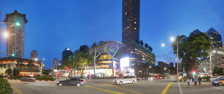 Panoramic street view of Orchard Road in Singapore at twilight showing ION Orchard shopping mall with curved glass canopy, illuminated buildings, cars on the road, pedestrians crossing, and tropical trees under deep blue evening sky
