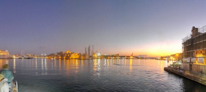 Panoramic view of Valletta's Grand Harbour at dusk showing illuminated historic buildings, industrial cranes, and water reflections under a purple-blue twilight sky