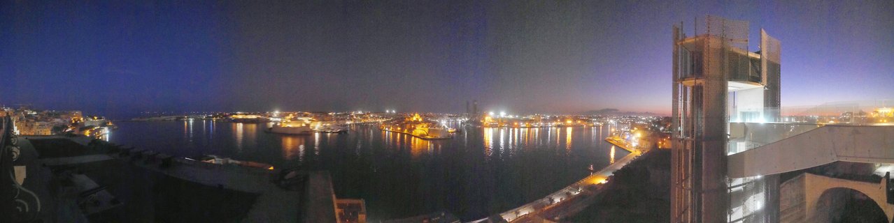 Panoramic night view of Valletta Grand Harbour showing illuminated historic fortifications, city lights reflecting on water, and modern buildings under deep blue twilight sky