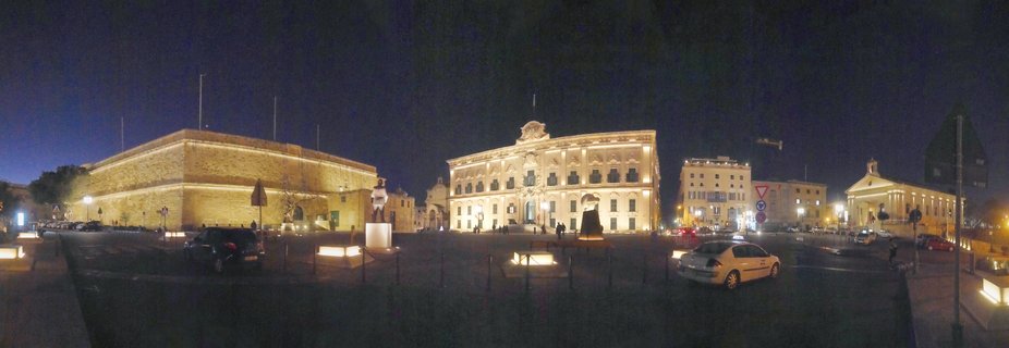 Panoramic night photograph of illuminated historic baroque buildings and stone fortifications in a public square in Valletta, Malta, with parked cars and pedestrians visible