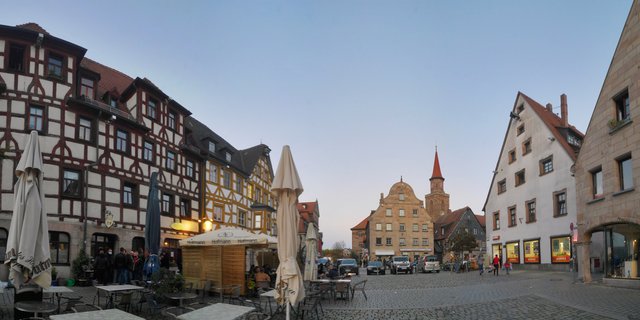 Historic German town square with half-timbered buildings, cobblestone pavement, outdoor café seating with umbrellas, pedestrians, and a church tower with red spire in the background at dusk