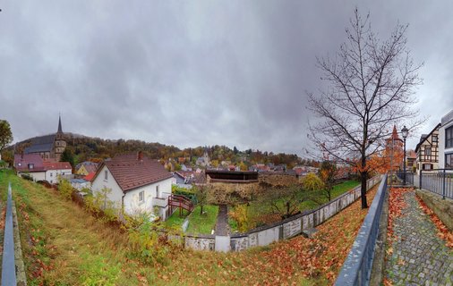 Panoramic view of Kulmbach, Germany showing medieval church spires, traditional Bavarian houses with red tile roofs, cobblestone path, stone walls, bare trees, and autumn leaves under cloudy sky