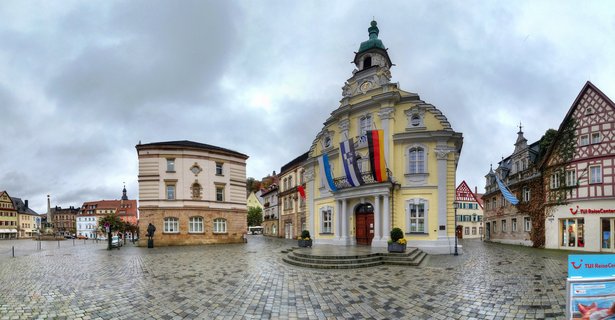 Panoramic view of Kulmbach's market square showing a yellow baroque town hall with green dome, German and Bavarian flags, cobblestone pavement, and traditional half-timbered buildings under cloudy sky