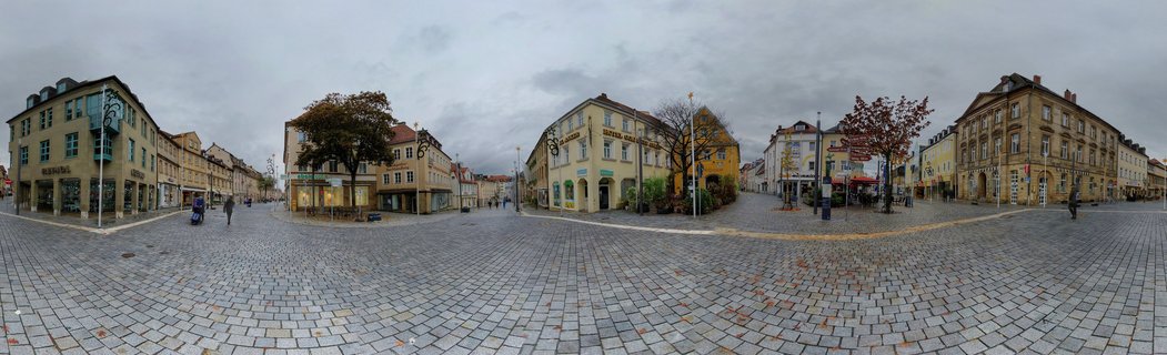 Panoramic street view of a German town square with cobblestone pavement, multi-story historic buildings, autumn trees, and pedestrians under cloudy sky